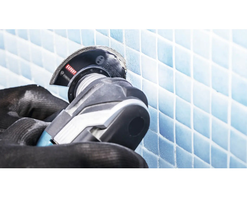 Une paire de mains gantées utilise un outil multifonction Bosch avec une lame diamantée pour couper des carreaux de mosaïque.