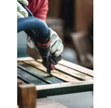 Person mit Arbeitshandschuhen verwendet ein Multifunktionswerkzeug mit Absaugvorrichtung, um eine Holzoberfläche zu schleifen.