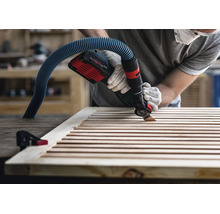Handwerker mit Arbeitshandschuhen verwendet ein Akku-Multitool mit angeschlossener Staubabsaugung, um Holzlamellen zu bearbeiten.