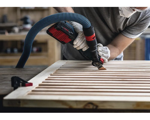 Handwerker mit Arbeitshandschuhen verwendet ein Akku-Multitool mit angeschlossener Staubabsaugung, um Holzlamellen zu bearbeiten.