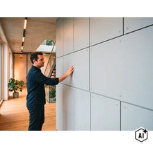Un homme touche un revêtement mural composé de panneaux carrés dans un espace de vie moderne avec parquet et plafond en béton apparent. (Image AI modified or generated)
