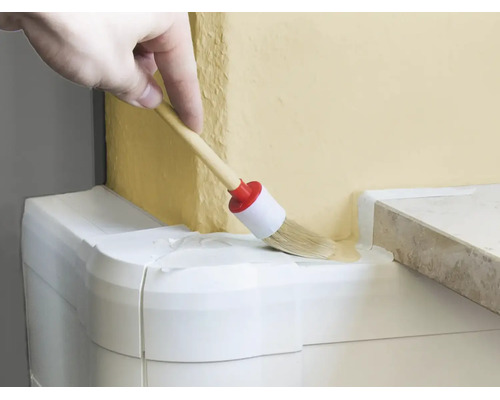 Eine Hand nutzt einen Rundpinsel, um eine Wand an einem Übergang zu einer weissen Leiste zu streichen.