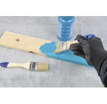 Person streicht mit einem Pinsel eine Holzplatte mit blauer Farbe, eine Farbdose und ein weiterer Pinsel liegen daneben.