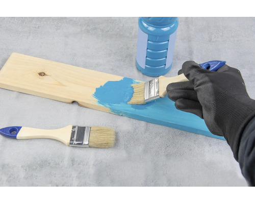 Person streicht mit einem Pinsel eine Holzplatte mit blauer Farbe, eine Farbdose und ein weiterer Pinsel liegen daneben.