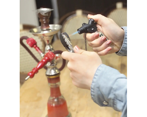 Une main allume du charbon à chicha avec un chalumeau à gaz, une chicha est posée sur une table en arrière-plan.