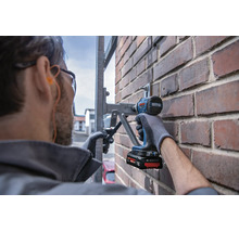 Artisan avec lunettes de protection fixe un support métallique sur un mur en briques avec une visseuse à chocs sans fil Bosch.