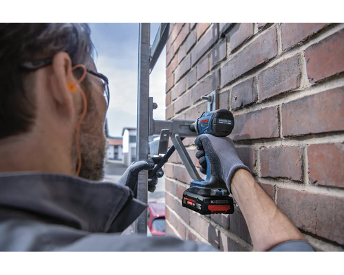 Artisan avec lunettes de protection fixe un support métallique sur un mur en briques avec une visseuse à chocs sans fil Bosch.