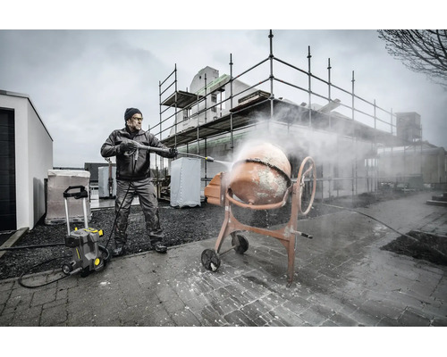 Un homme nettoie une bétonnière avec un nettoyeur haute pression sur un chantier devant une maison avec échafaudage.