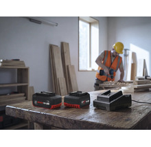 Zwei Bosch 18 Volt Akkus und ein Ladegeraet stehen auf einer Werkbank, waehrend im Hintergrund ein Handwerker mit einer Bohrmaschine arbeitet.