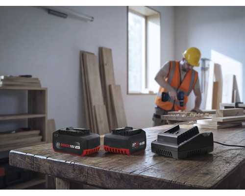 Zwei Bosch 18 Volt Akkus und ein Ladegeraet stehen auf einer Werkbank, waehrend im Hintergrund ein Handwerker mit einer Bohrmaschine arbeitet.