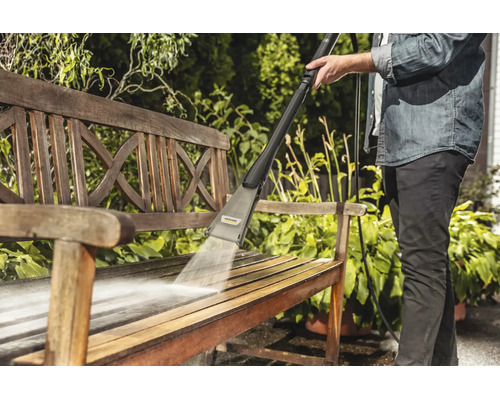 Une personne nettoie un banc de jardin en bois avec un nettoyeur haute pression Karcher en extérieur.