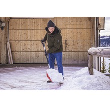 Eine Person schaufelt Schnee mit einer Schneeschaufel vor einem Holzgebäude.