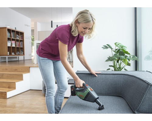 Une femme aspire un canapé gris avec un aspirateur sans fil dans un salon lumineux.