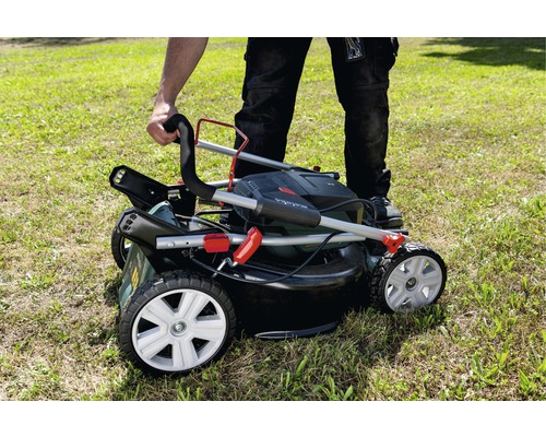 Une personne plie une tondeuse à gazon avec bac de ramassage sur une pelouse.