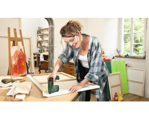 Frau spannt Leinwand mit einem elektrischen Tacker in einem hellen Atelier