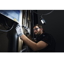 Un homme portant des lunettes de protection et des gants travaille avec une visseuse sans fil sur un objet, éclairé par une lampe de travail.