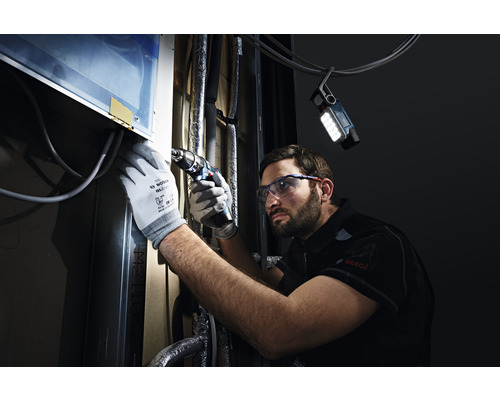 Un homme portant des lunettes de protection et des gants travaille avec une visseuse sans fil sur un objet, éclairé par une lampe de travail.