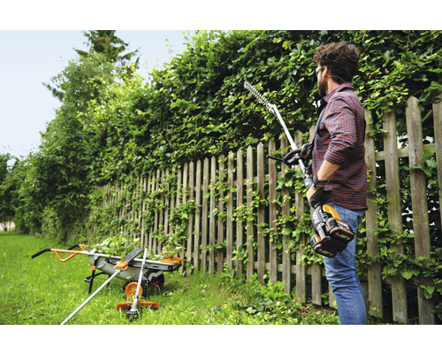 Un homme taille une haie avec un taille-haie à batterie dans le jardin. À l''arrière-plan, on aperçoit une brouette et un coupe-bordures.