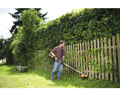 Un homme coupe l''herbe avec un coupe-bordures sans fil dans un jardin avec une clôture en bois et une haie.