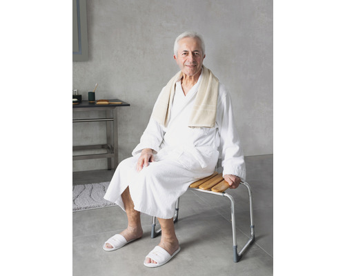 Un homme en peignoir est assis sur un tabouret de douche avec des lattes en bois et un cadre en métal.