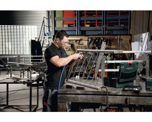 Un homme travaille un garde-corps en métal avec un outil pneumatique dans un atelier. Une boîte à outils de Metabo est posée sur la table.