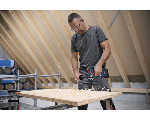 Un homme perce un trou dans une planche de bois avec une perceuse-visseuse sans fil dans un grenier.