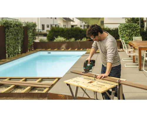 Un homme coupe une planche en bois avec une scie sauteuse dans le jardin, une piscine en arrière-plan