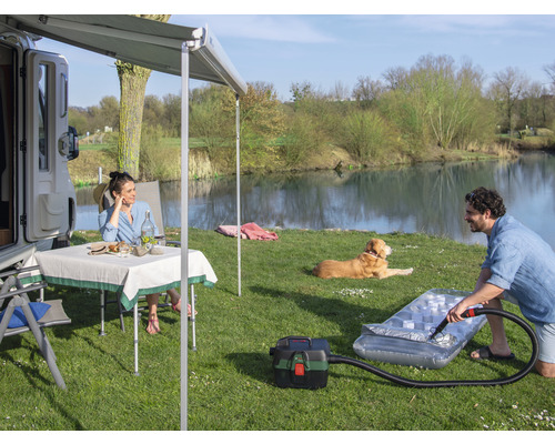 Scène avec camping-car, store, table, chaises, chien et personne gonflant un matelas pneumatique avec un aspirateur, à l'extérieur au bord d'un lac.