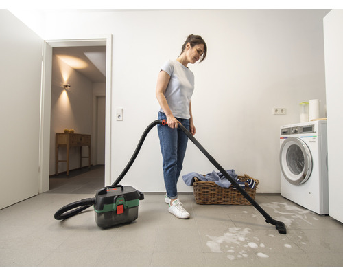 Une femme passe l'aspirateur avec un aspirateur polyvalent dans une buanderie. À l'arrière-plan, il y a une machine à laver et un panier à linge.