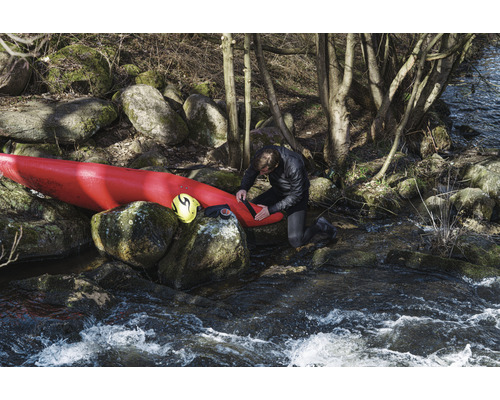 Mann repariert Kajak am Flussufer mit Felsen und Bäumen.