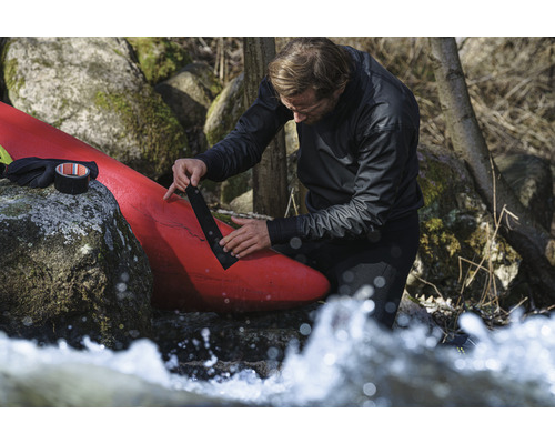 Ein Mann repariert ein rotes Kajak mit schwarzem Panzertape in der Nähe von Felsen und fließendem Wasser.