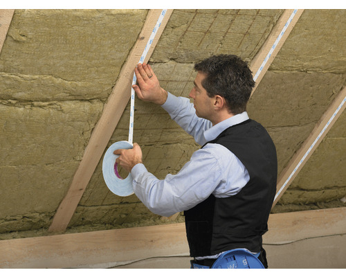 Un homme fixe du ruban adhésif double face sur une latte en bois dans un grenier isolé.