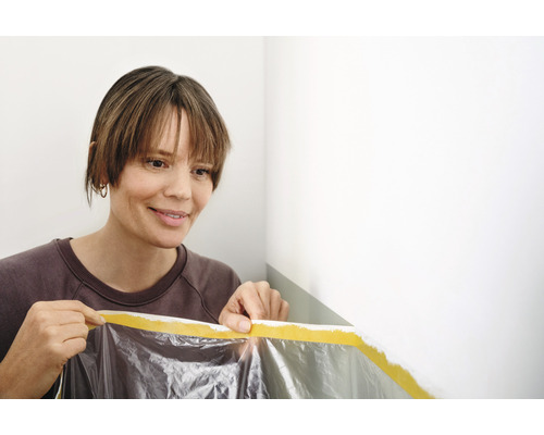 Frau befestigt Abdeckfolie mit Klebeband an der Wand zum Malen