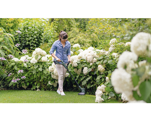 Une femme utilise un coupe-bordures dans un jardin avec des arbustes en fleurs.