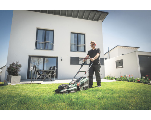 Un homme tond la pelouse devant une maison moderne avec une tondeuse à gazon sans fil.