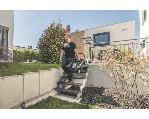Un homme porte une tondeuse à gazon pliée dans un escalier du jardin