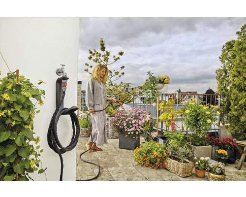 Frau bewässert Pflanzen auf dem Balkon mit einem Gartenschlauch, der an einer Wandhalterung befestigt ist