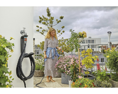 Une femme arrose des plantes sur un balcon avec un tuyau d''arrosage fixé à un support mural.