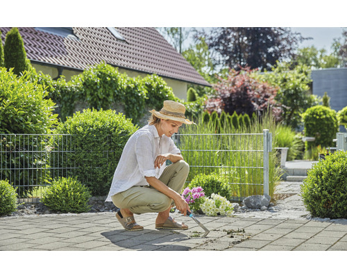 Frau entfernt Unkraut mit einem Fugenkratzer auf einer gepflasterten Terrasse.