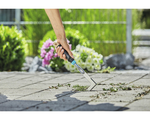 Eine Person entfernt Unkraut mit einem Fugenkratzer von Pflastersteinen in einem Garten.