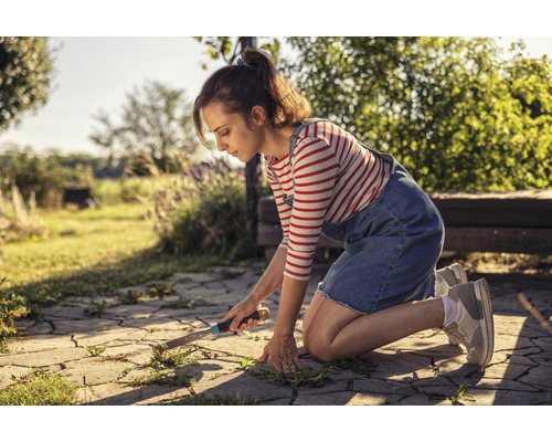 Frau entfernt Unkraut von Pflastersteinen im Garten mit einem Unkrautstecher.