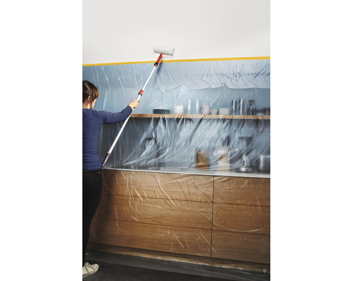 Une femme peint un mur avec un rouleau dans une cuisine recouverte de film plastique. Travaux de peinture pour la rénovation.