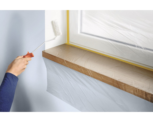 Une personne peint un mur avec un rouleau à peinture. La fenêtre et le rebord de la fenêtre sont recouverts de film plastique et de ruban adhésif.