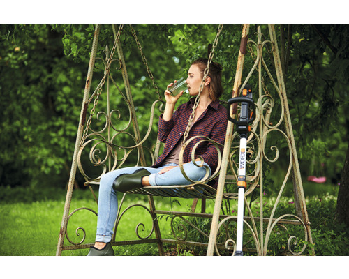 Une femme est assise sur une balançoire de jardin à côté d''un coupe-bordures WORX et boit une boisson.