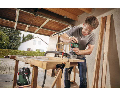 Mann arbeitet mit einer Oberfräse in einer Werkstatt an einem Holztisch. Ein Staubsauger steht daneben.