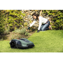 Ein Rasenmähroboter steht auf einer Rasenfläche, während eine Frau im Hintergrund im Garten arbeitet.