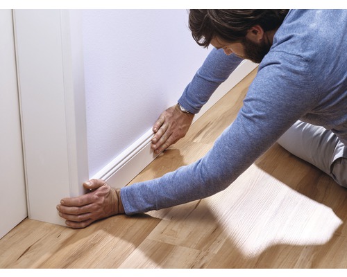 Un homme installe une plinthe blanche sur un mur dans une pièce avec un plancher en bois.