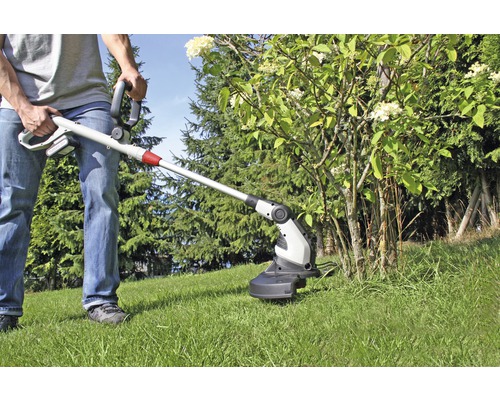 Eine Person benutzt einen Akku-Rasentrimmer im Garten, um das Gras unter einem Strauch zu schneiden.