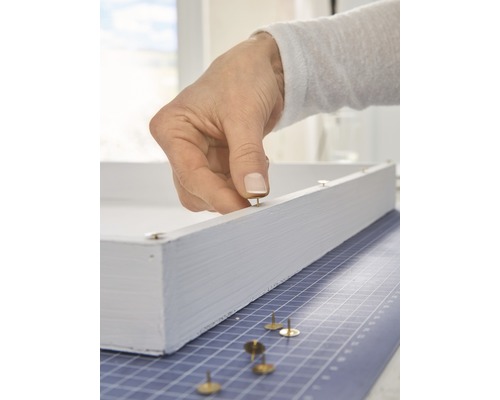 Une main fixe une punaise sur un cadre en bois blanc.