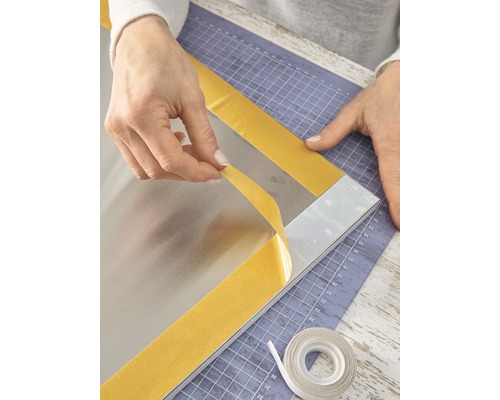 Une personne fixe un miroir avec du ruban adhésif double face sur un tapis de découpe.